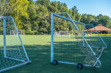 Arka planda bir futbol takımıyla birlikte büyük ağların arasından topu sahadan atarak spor parkındaki köşkten uzağa doğru kovalamaya çalışıyor.