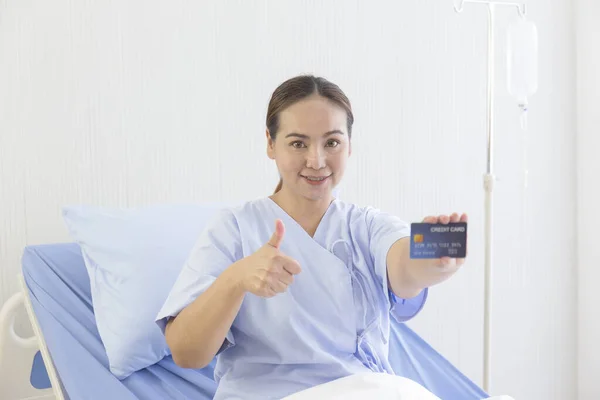 An Asian female patient lay on the hospital bed in her hand, held a credit card, and thumbs up to show a smile.