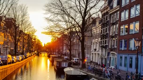 日落映照在阿姆斯特丹运河的水中 背景是美丽的房屋 树木与停放的船只 自行车和汽车与教堂的轮廓在远处 时间推移 — 图库视频影像