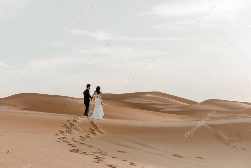 A couple in their wedding dresses walk barefoot through the desert at dawn. Wedding concept
