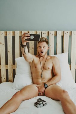 Yatakta çıplak genç bir adam takipçileri için selfie çekiyor. Yaşam tarzı kavramı
