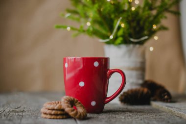 Eski ahşap masa (arka plan) üzerinde beyaz benekli kırmızı kupa. Arka planda, ışık çelenkleri olan bir Noel ağacı.
