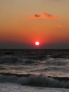 Koyu fırtınalı denizle kumsalda güzel, canlı, kırmızı bir gün batımı. Beyaz köpük ve bulutlarla kaplanmış dramatik bir manzara.