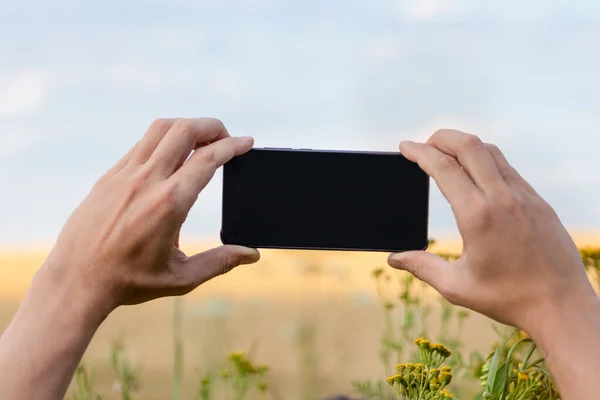 Holding smart phone with black screen with two hands horizontally in sunny flowers field with blue sky natural landscape mockup template