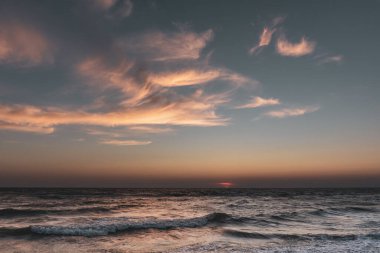 Gün batımı ufukta gizlenmiş fırtınalı dalgalarla deniz kıyıları ve sıcak güneş ışınlarıyla aydınlanan destansı renkli bulutlar.