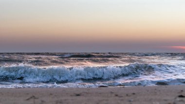 Fırtınalı beyaz dalgalar deniz kıyısındaki kumsalda yakın plan güneş batarken gökyüzü pastel renklerle