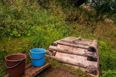 Ahşap kuyusu ve yeşil çimenleri olan bir yamaçta kovalarca suyla kırsal geçmişe ait manzaralar.