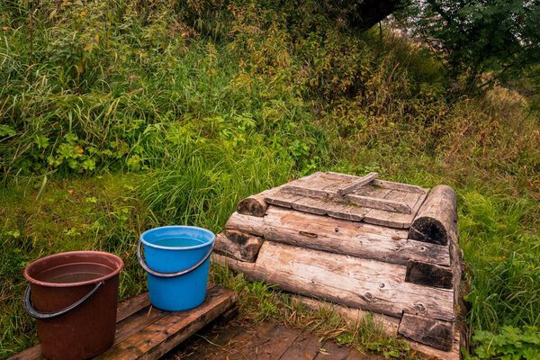 Сельский ретро-ландшафт с деревянным колодцем и ведрами воды на склоне холма с зеленой травой