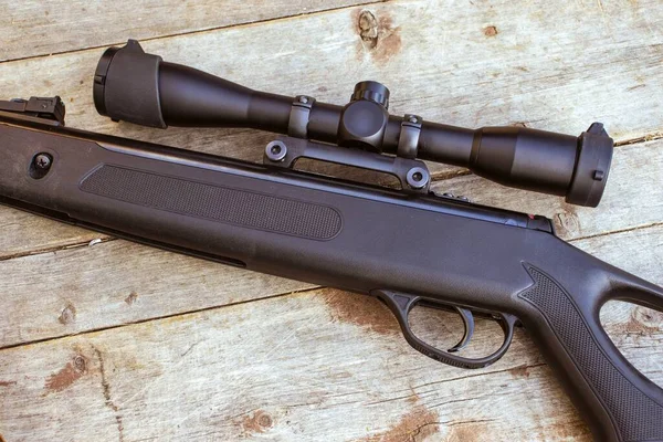 Part of a black rifle with a telescopic sight on a wooden background. Close-up