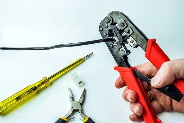 Hand of a craftsman with a crimping tool connecting an RJ45 connector with a black cable on a white background with a screwdriver and pliers. Networks and telecommunications