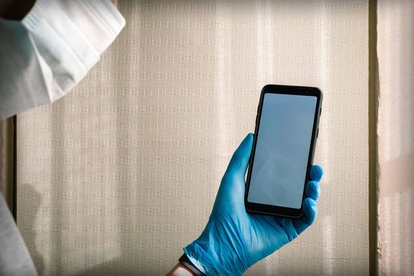 A man in a medical mask holds a smartphone with a white blank screen in his hand in a blue medical glove. Protection against bacteria in quarantine for coronavirus.