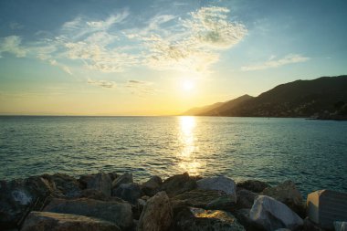 Sakin denizin üzerinde altın gün batımı, ufukta kayalık kıyı ve dağlar..