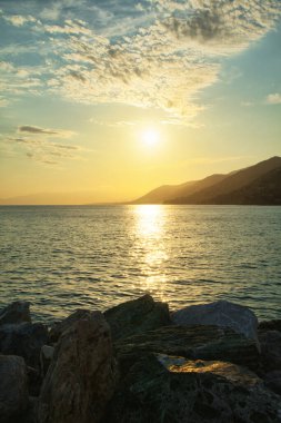 Sakin denizin üzerinde altın gün batımı, ufukta kayalık kıyı ve dağlar..