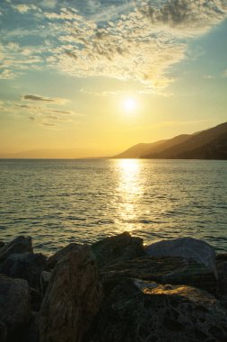 Sakin denizin üzerinde altın gün batımı, ufukta kayalık kıyı ve dağlar..
