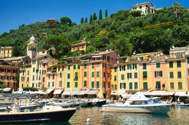 Colorful waterfront houses and boats in the picturesque harbor of Portofino, Liguria, Italy, with the church tower and green hills in the background.