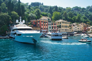 Colorful waterfront houses and boats in the picturesque harbor of Portofino, Liguria, Italy, with the church tower and green hills in the background.