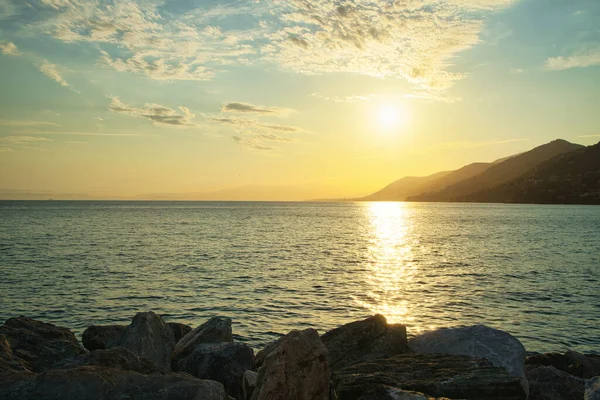 Sakin denizin üzerinde altın gün batımı, ufukta kayalık kıyı ve dağlar..
