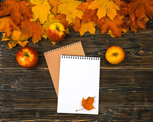 Um caderno com um espaço vazio para texto, maçãs em uma pilha de folhas de outono brilhantes contra uma mesa de madeira escura — Fotografia de Stock