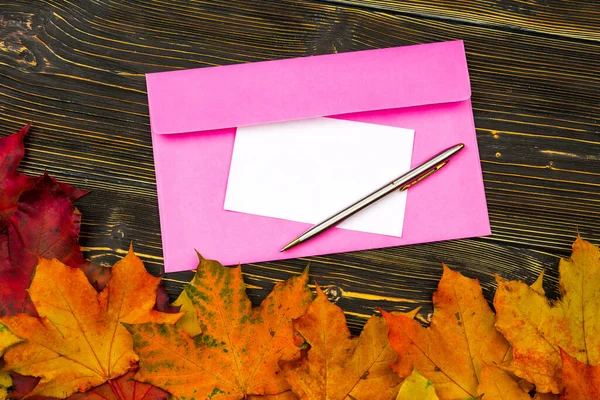 Een enveloppe en papier met een lege ruimte voor tekst op een stapel heldere herfstbladeren tegen een donkere houten tafel — Stockfoto