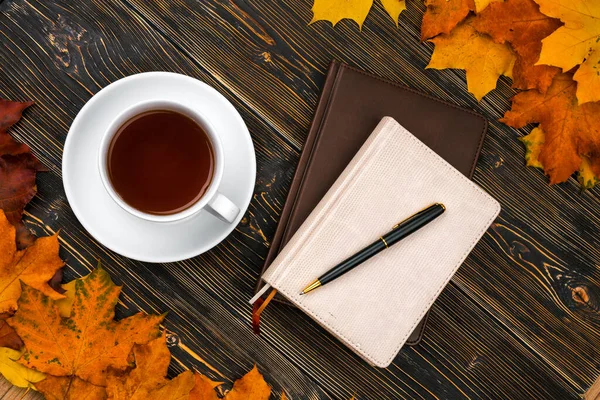 Notitieboek dagboek Kopje thee op een stapel heldere herfstbladeren tegen een donkere houten tafel — Stockfoto