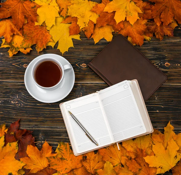 Un carnet avec un espace vide pour le texte, une tasse de thé sur une pile de feuilles d'automne lumineux contre une table en bois sombre Photos De Stock Libres De Droits