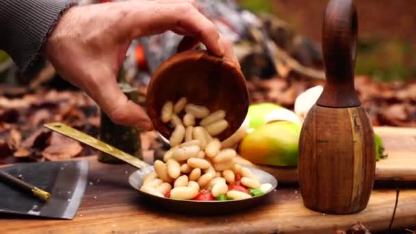 キャンプファイヤーの木板に缶詰のハリコット豆を新鮮なカット野菜ボウルサラダトマトペッパー玉ねぎに入れる男の手 — ストック動画
