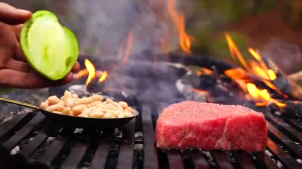 男の手は 森林キャンプ火災の炎にビーフステーキピンクの肉の大きな部分と黒いグリルパンに新鮮なカット野菜を置きます — ストック動画