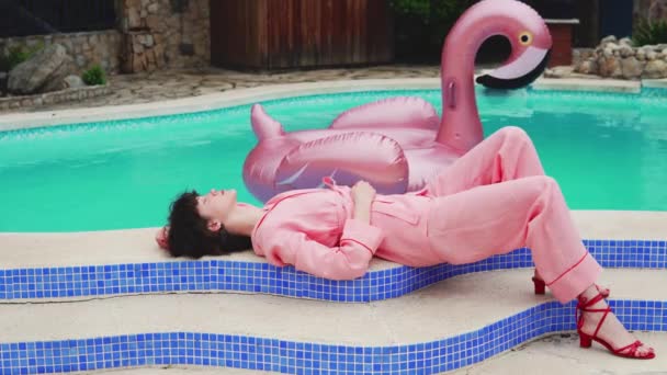 ピンクのジャムの若いモデルの女性は、フローティングピンクのフラミンゴとプールの近くに位置しています — ストック動画