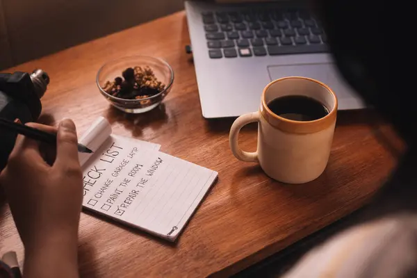 Bir kadının eli, bir fincan kahve, fındık, angarya bir liste, bir dizüstü bilgisayar ve aletlerle ev eşyaları hazırlıyor.