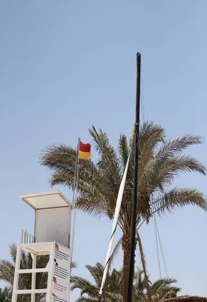 Rescue tower with red and orange flag on the beach against a palm tree