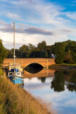A typical Suffolk scene of a sail boat moored up on the rivers edge with a brick bridge in the background with the reflection in the river below clipart