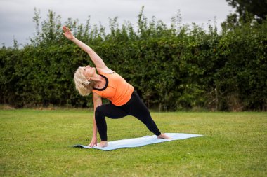 Çimenli bir parkta çıplak ayak yoga yapan orta yaşlı bir kadın.