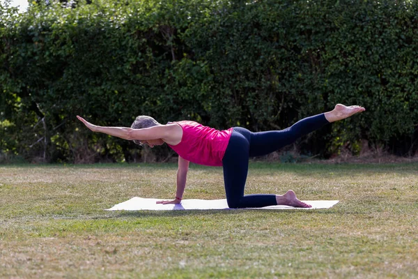 A middle aged woman practicing yoga barefoot outside in a grassy