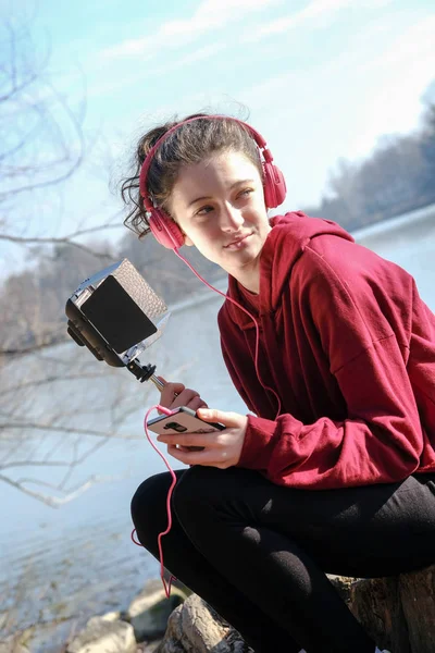 smiling teenager girl with pink headphoens listen to music on a mobile outside near a lake