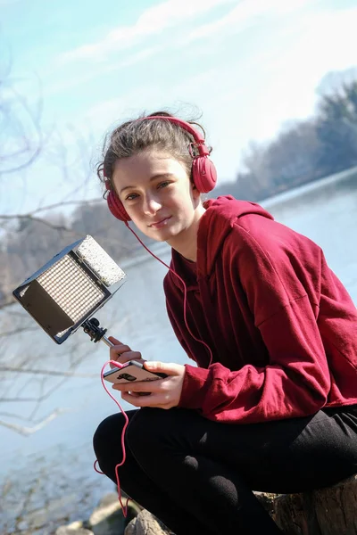 smiling teenager girl with pink headphoens listen to music on a mobile outside near a lake