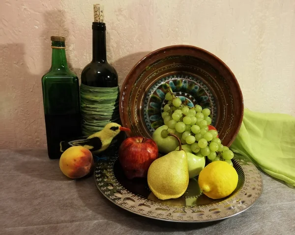 Masanın üzerinde yuvarlak bir metal tepsi var. Tepside meyveler vardır: armut, limon, elma ve yeşil üzüm. Tepsinin solunda bir şeftali, iki şişe şarap ve sarı bir kuş vardı. Tepsinin arkasında büyük bir kil çanak. Bej arka plan.