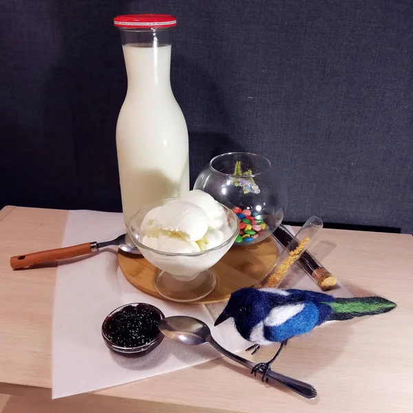 On the table is a round wooden stand. On it is a bottle of milk, a bowl of ice cream, a round vessel with colored jelly beans and a spoon. Next to a white napkin is a small bowl of jam, bird, metal spoon and the two vials. Background dark blue.