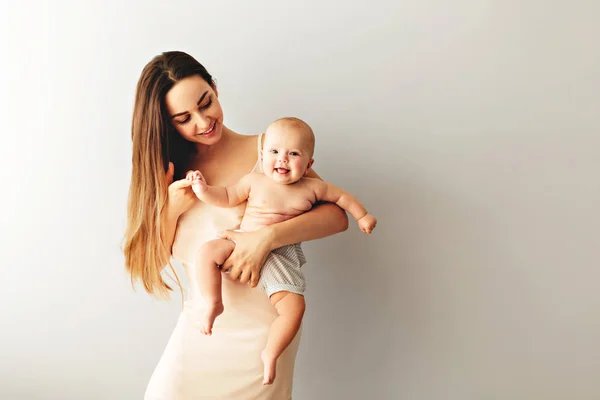 Bela mãe segura seu bebê em um fundo claro simples . — Fotografia de Stock