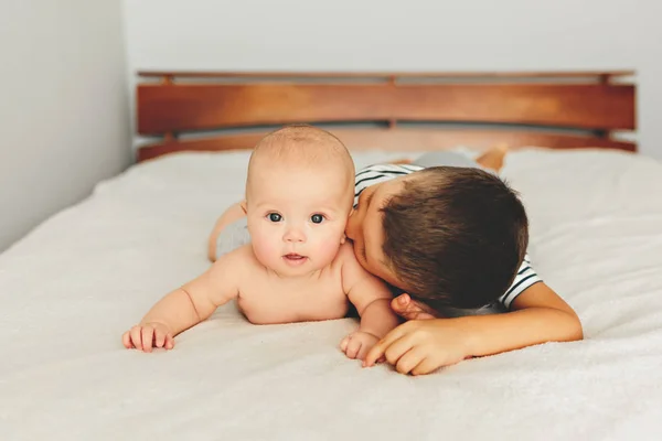 Lindo bebê jaz na cama com seu irmão mais velho. A criança beija sua irmãzinha . — Fotografia de Stock
