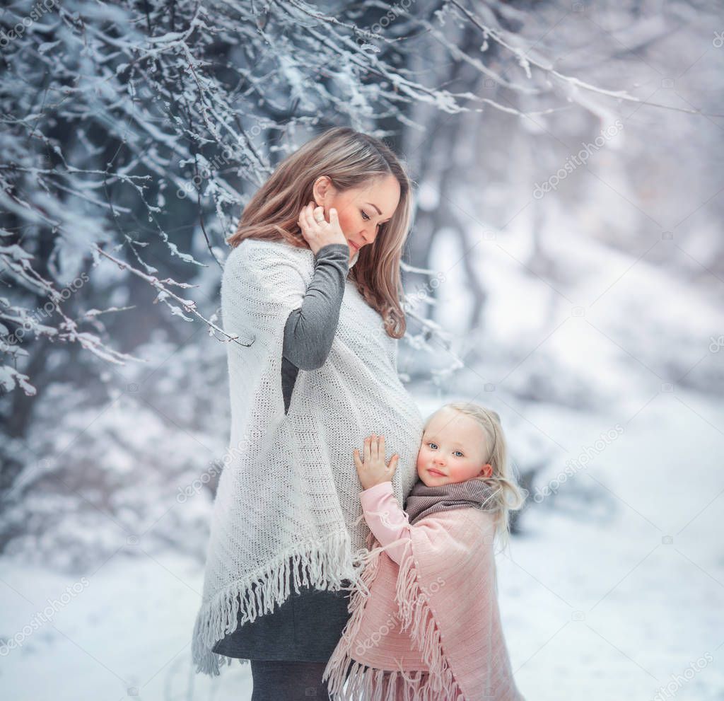 young beautiful pregnant mother with her daughter in a snowy forest in a delicate pink sweater. girl listening to belly