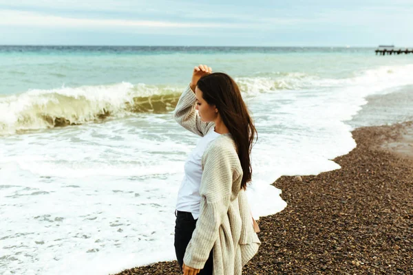 Una hermosa joven camina a lo largo de la orilla del mar, una tormenta, el pelo vuela aparte, un cárdigan gris, una figura deportiva en zapatillas de deporte disfruta, otoño o invierno — Foto de Stock