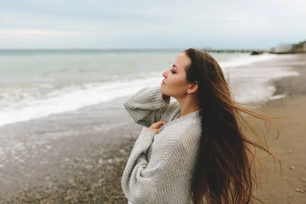 Una hermosa joven camina a lo largo de la orilla del mar, una tormenta, el pelo vuela aparte, un cárdigan gris, una figura deportiva en zapatillas de deporte disfruta, otoño o invierno — Foto de Stock