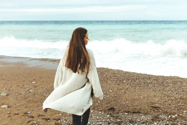 Una hermosa joven camina a lo largo de la orilla del mar, una tormenta, el pelo vuela aparte, un cárdigan gris, una figura deportiva en zapatillas de deporte disfruta, otoño o invierno — Foto de Stock