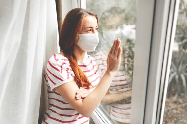 Salgın konsepti. Karantinada tıbbi maskeli üzgün genç bir kadının portresi, pencerenin yanındaki bir hastanede yatan kız hasta.