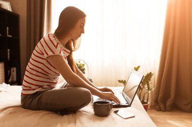 Güzel bir genç kadının dizüstü bilgisayarla çalışırken cep telefonunu kullanırken evde otururken, karantinaya alınırken görüntüsü.