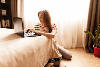 Güzel bir genç kadının dizüstü bilgisayarla çalışırken cep telefonunu kullanırken evde otururken, karantinaya alınırken görüntüsü.