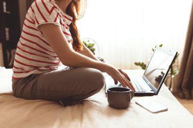 Güzel bir genç kadının dizüstü bilgisayarla çalışırken cep telefonunu kullanırken evde otururken, karantinaya alınırken görüntüsü.