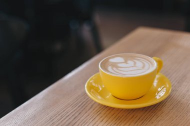 Bir fincan kapuçino, kafedeki ahşap bir masada latte sanatı. Sarı seramik bardak.