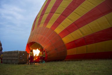 Crews ready hot air balloons before taking to flight during a cloudy morning over the savanna of the Masai Mara National Reserve, Kenya, Africa. Balloons inflating with hot air produced by flames.