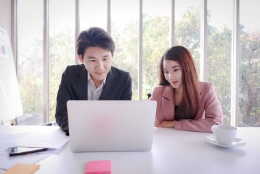 Genç güzel gülümseme Asyalı iş kadını ile genç Asya iş adamı ofiste dizüstü bilgisayar ile çalışır. Sezonluk satışların performansı hakkında tartışma. Belgeler, beyaz masa üzerinde beyaz fincan ve cep telefonu